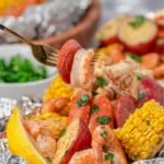 A fork holds shrimp and sausage above foil packets with corn, potatoes, shrimp, sausage, and a lemon wedge.