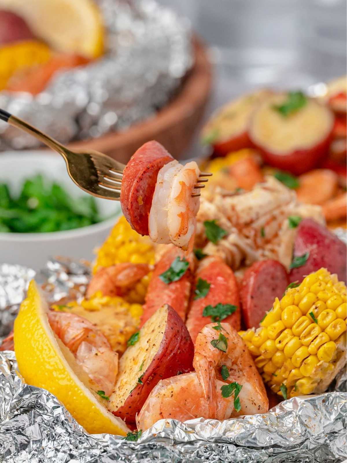 A fork holds shrimp and sausage above foil packets with corn, potatoes, shrimp, sausage, and a lemon wedge.