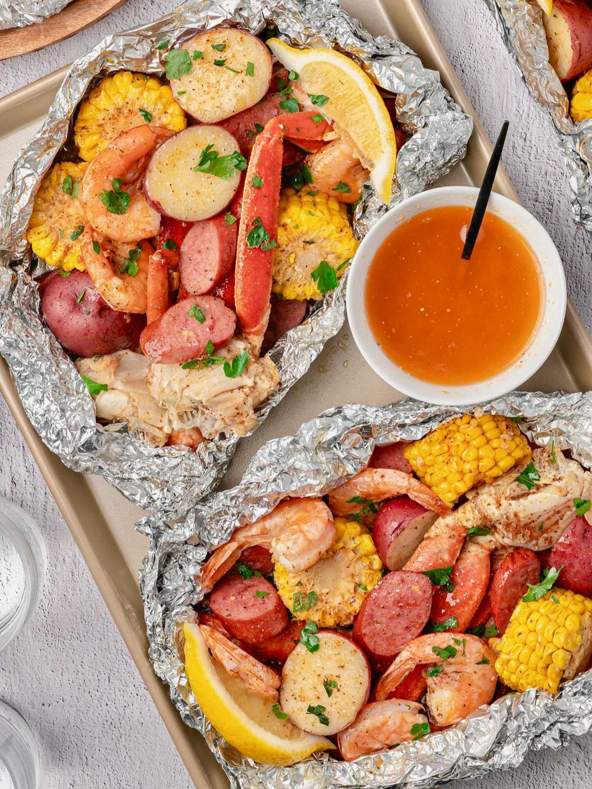 Foil packets with shrimp, sausage, corn, potatoes, and lemon slices, served with a side of dipping sauce.
