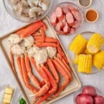 Raw crab legs, shrimp, sausage, corn, potatoes, spices, butter, lemon, garlic, and parsley on a white surface.
