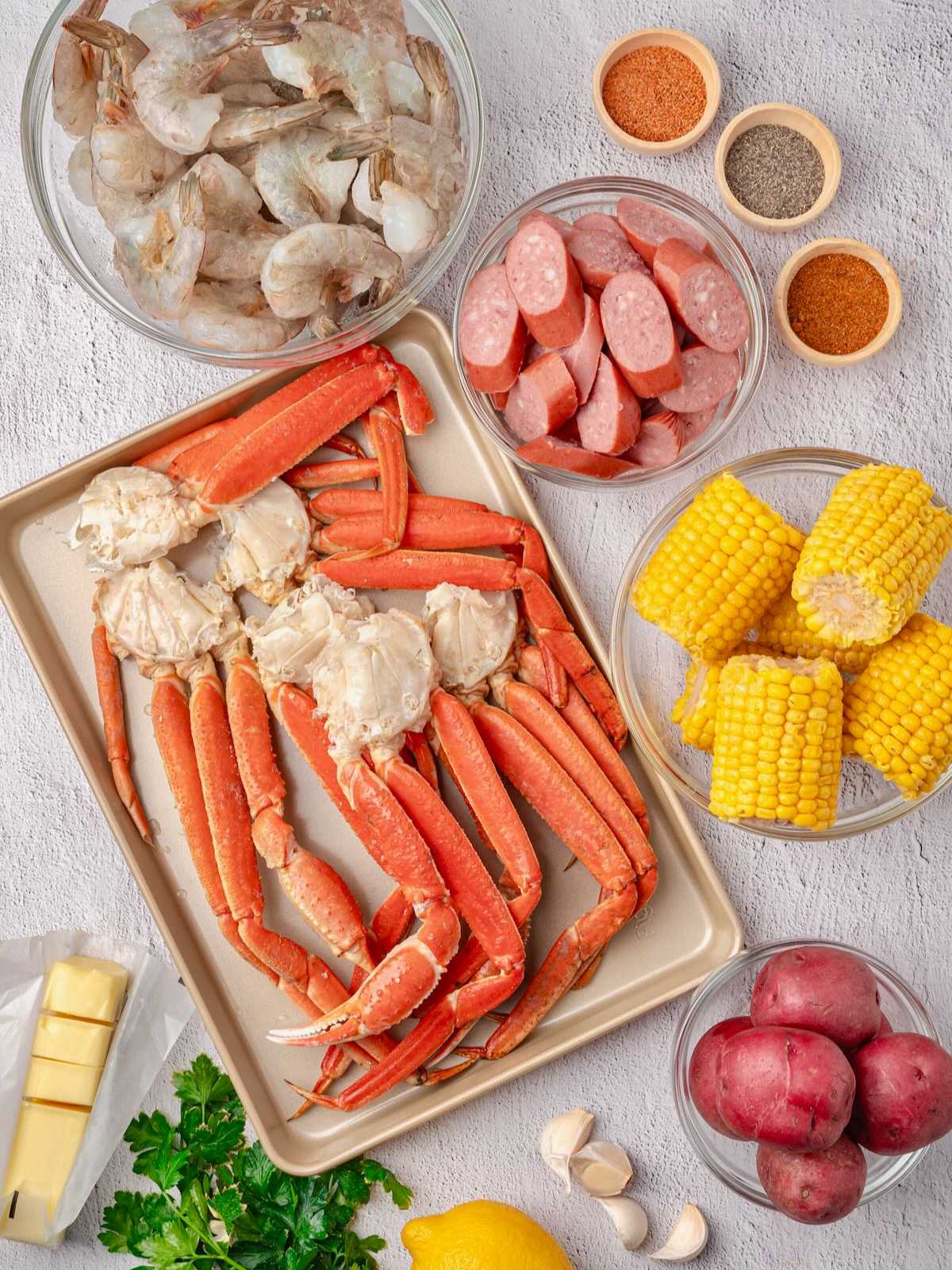 Raw crab legs, shrimp, sausage, corn, potatoes, spices, butter, lemon, garlic, and parsley on a white surface.