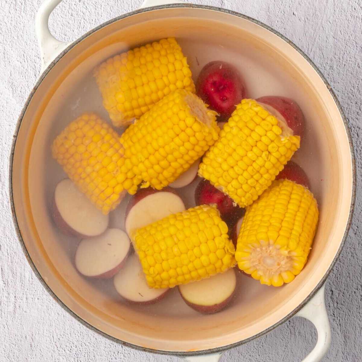 Corn on the cob and sliced red potatoes boiling in a large white pot filled with water.