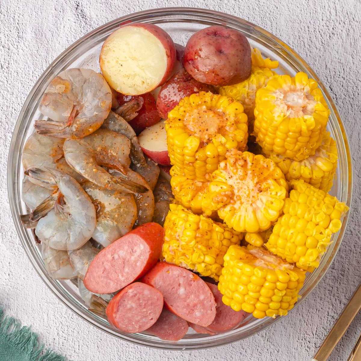 A bowl with raw shrimp, sliced sausage, corn on the cob pieces, and halved red potatoes.