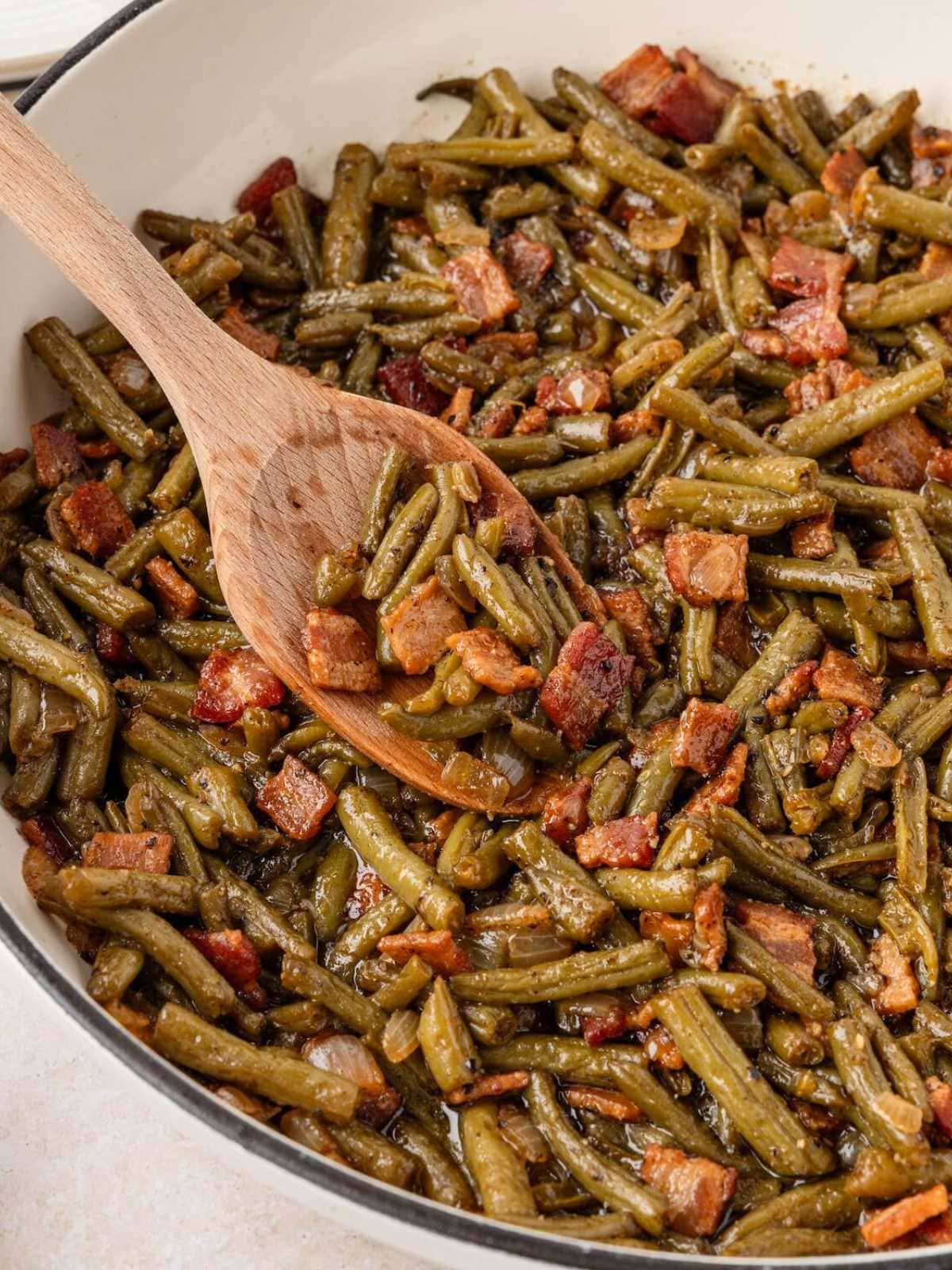 Smothered green beans with pieces of bacon being stirred in a pan with a wooden spoon.