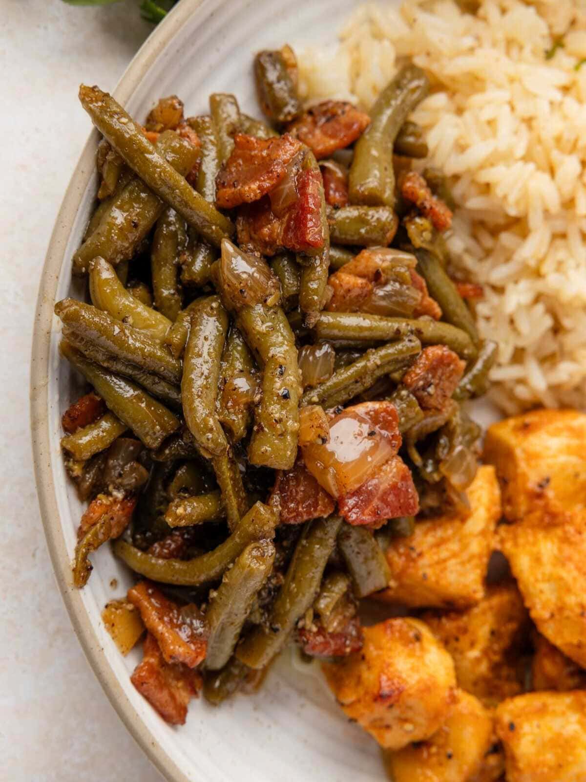 Green beans cooked with bacon and onions, served on a plate with rice and pieces of seasoned chicken.