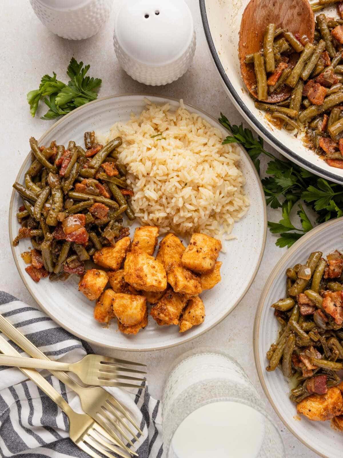 A plate with rice, green beans with bacon, and seasoned chicken pieces, next to a glass of milk.