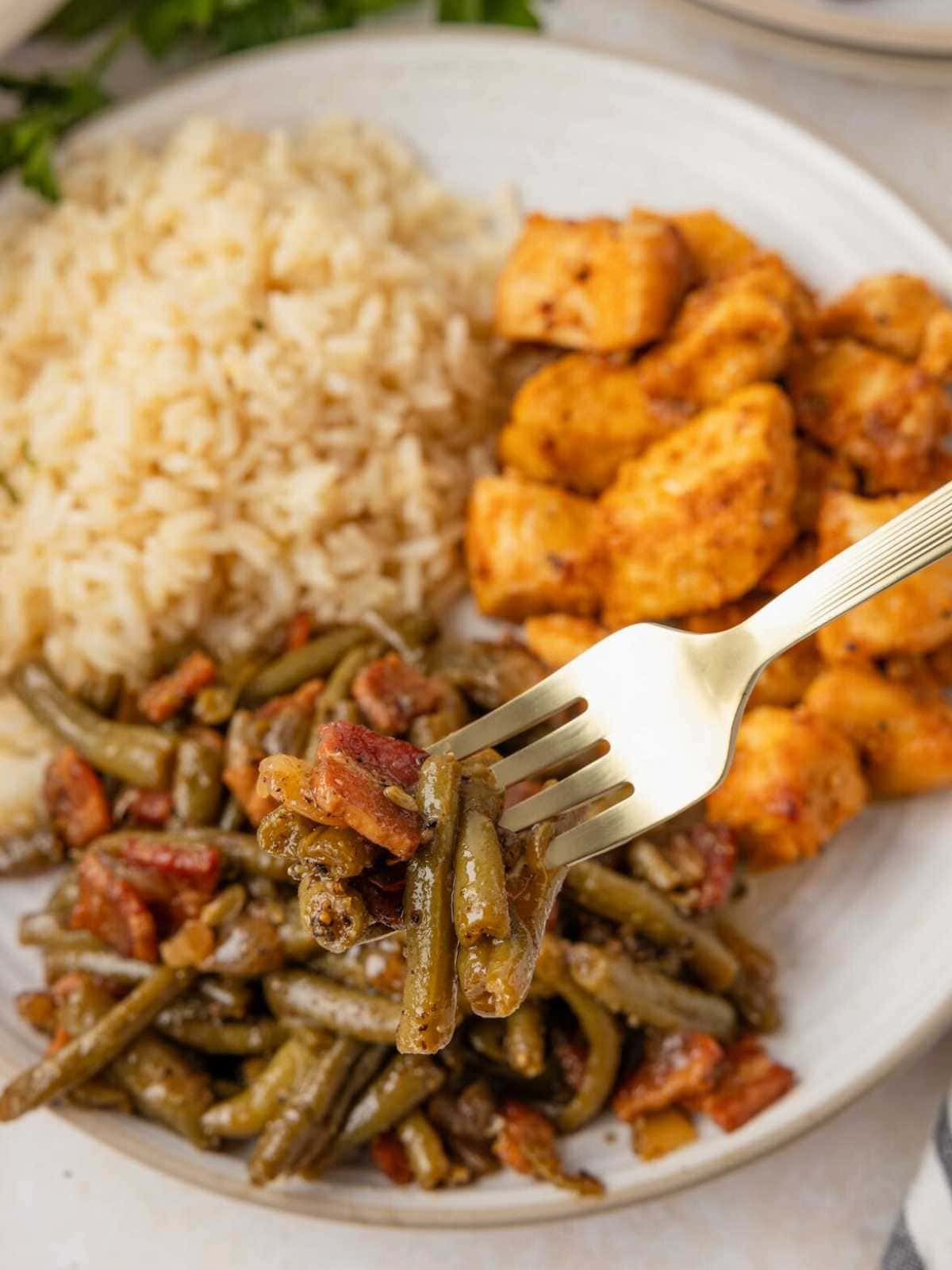 A fork holds green beans with bacon; rice and seasoned chicken are on a plate in the background.