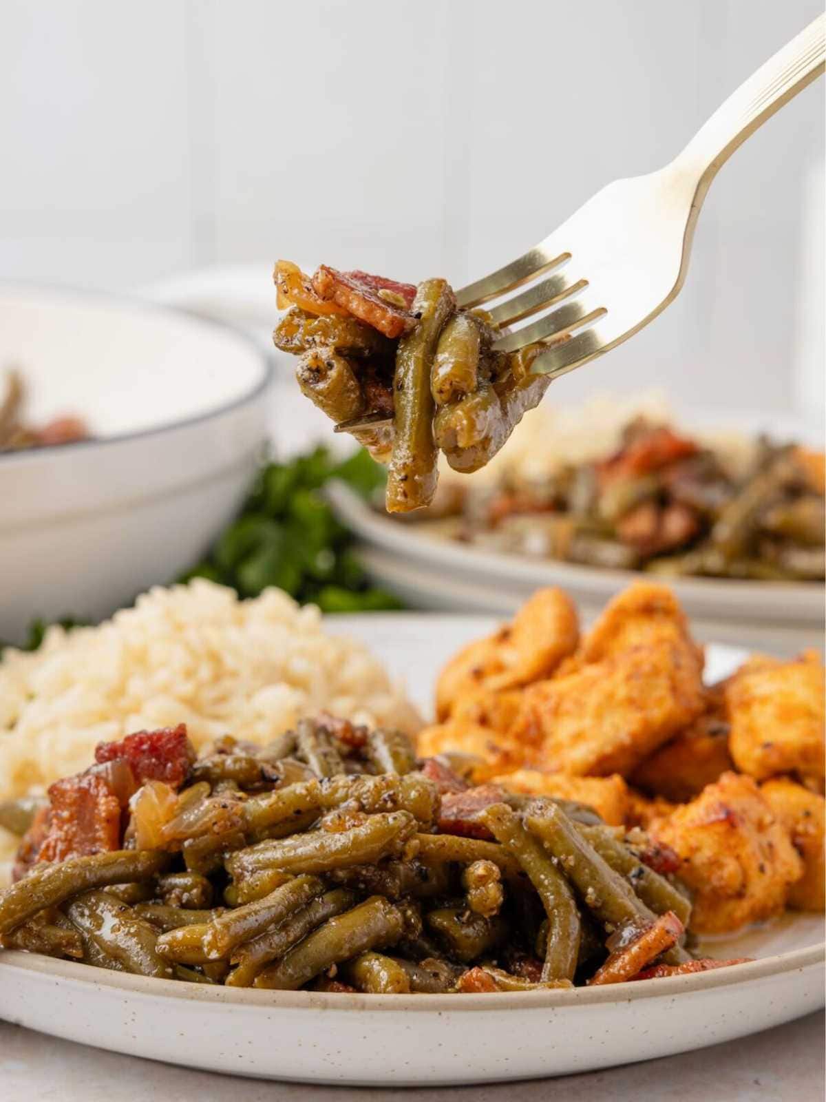 A fork holds green beans with bacon; rice and breaded chicken are served on a plate in the background.