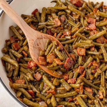 Cooked green beans with pieces of bacon in a pan, stirred with a wooden spoon.