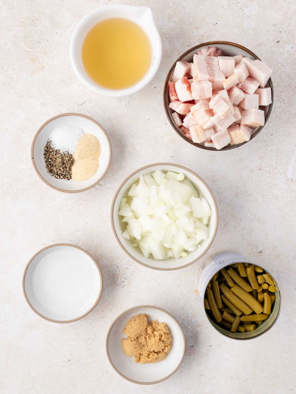 Seven bowls with chopped onions, cubed bacon, green beans, broth, spices, cream, and brown sugar on a light surface.