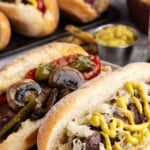 Two sausages in buns topped with mustard, sauerkraut, mushrooms, and peppers, with more food in the background.