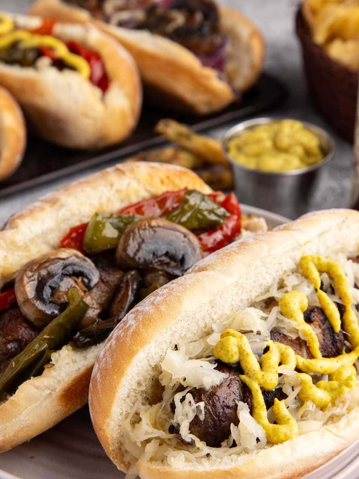 Two sausages in buns topped with mustard, sauerkraut, mushrooms, and peppers, with more food in the background.
