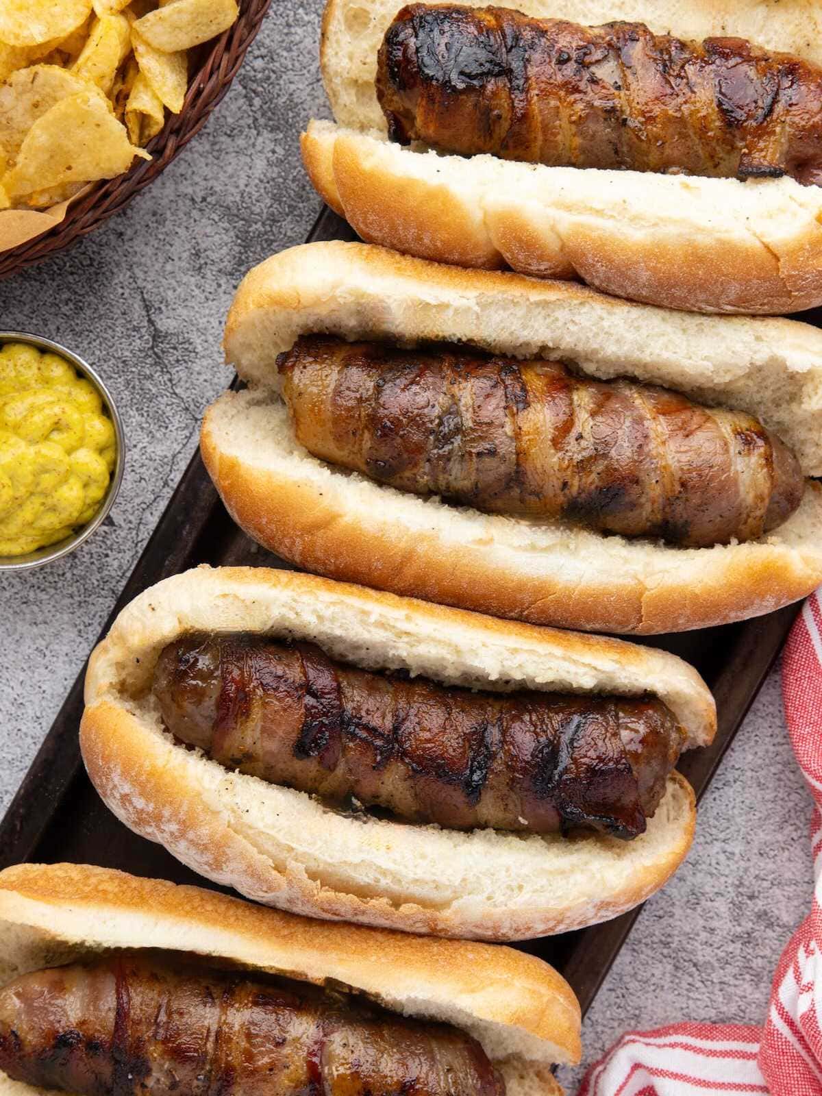 Four grilled sausages in buns on a tray, with chips and a bowl of yellow mustard nearby.