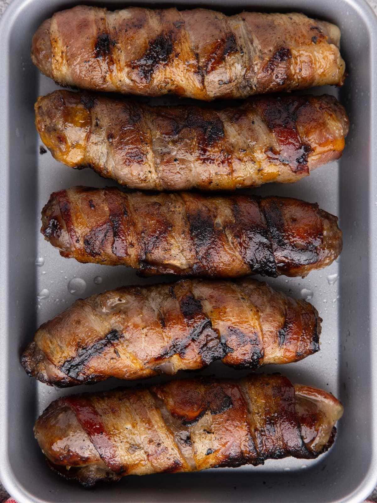 Five grilled bacon-wrapped sausages arranged in a baking pan.