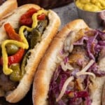 Two sausages in buns, one topped with peppers and mustard, the other with sautéed onions, with mustard in background.