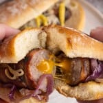 Hands hold a sausage sandwich with melted cheese, onions, and a soft bun; another sandwich is in the background.