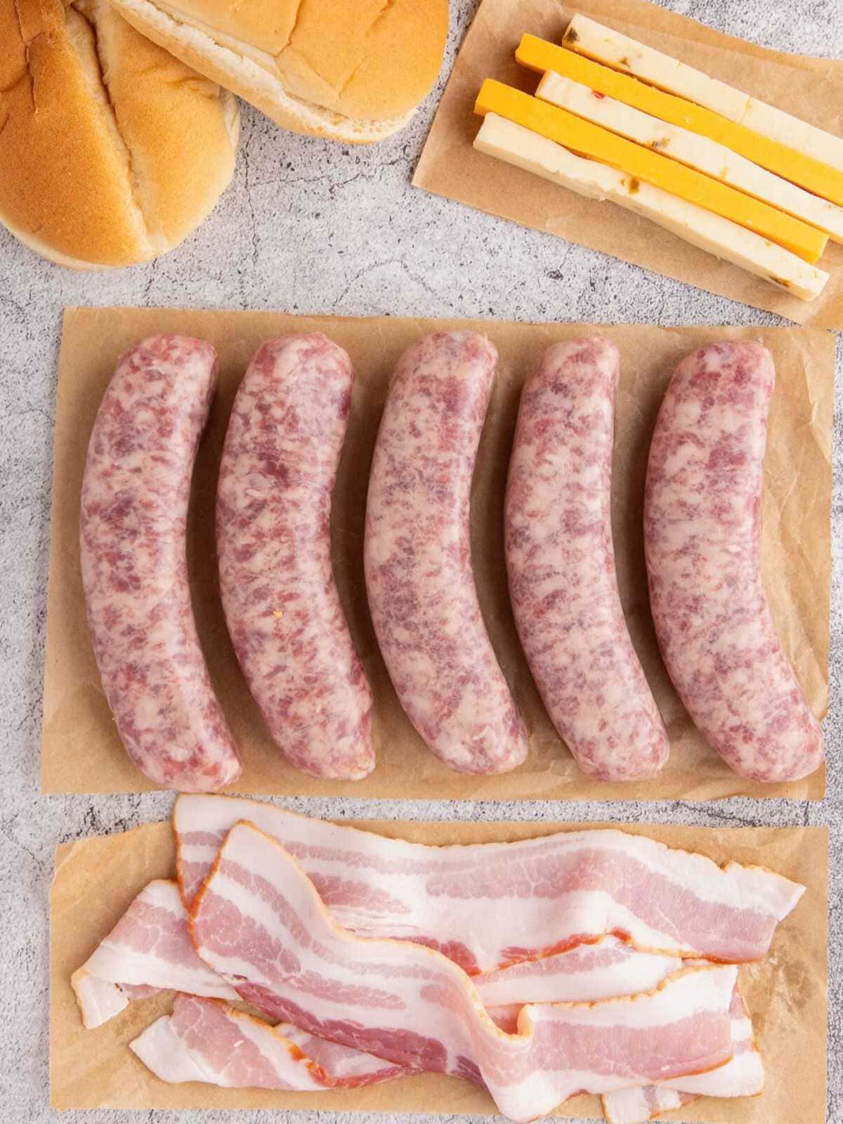 Raw bratwursts, bacon strips, cheddar cheese sticks, and sandwich buns on parchment paper.