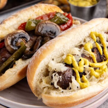 Two sausage sandwiches, one topped with mushrooms and peppers, the other with sauerkraut and yellow mustard.