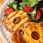 Grilled pork chops topped with pineapple rings, served with a green salad and cherry tomatoes on a white plate.