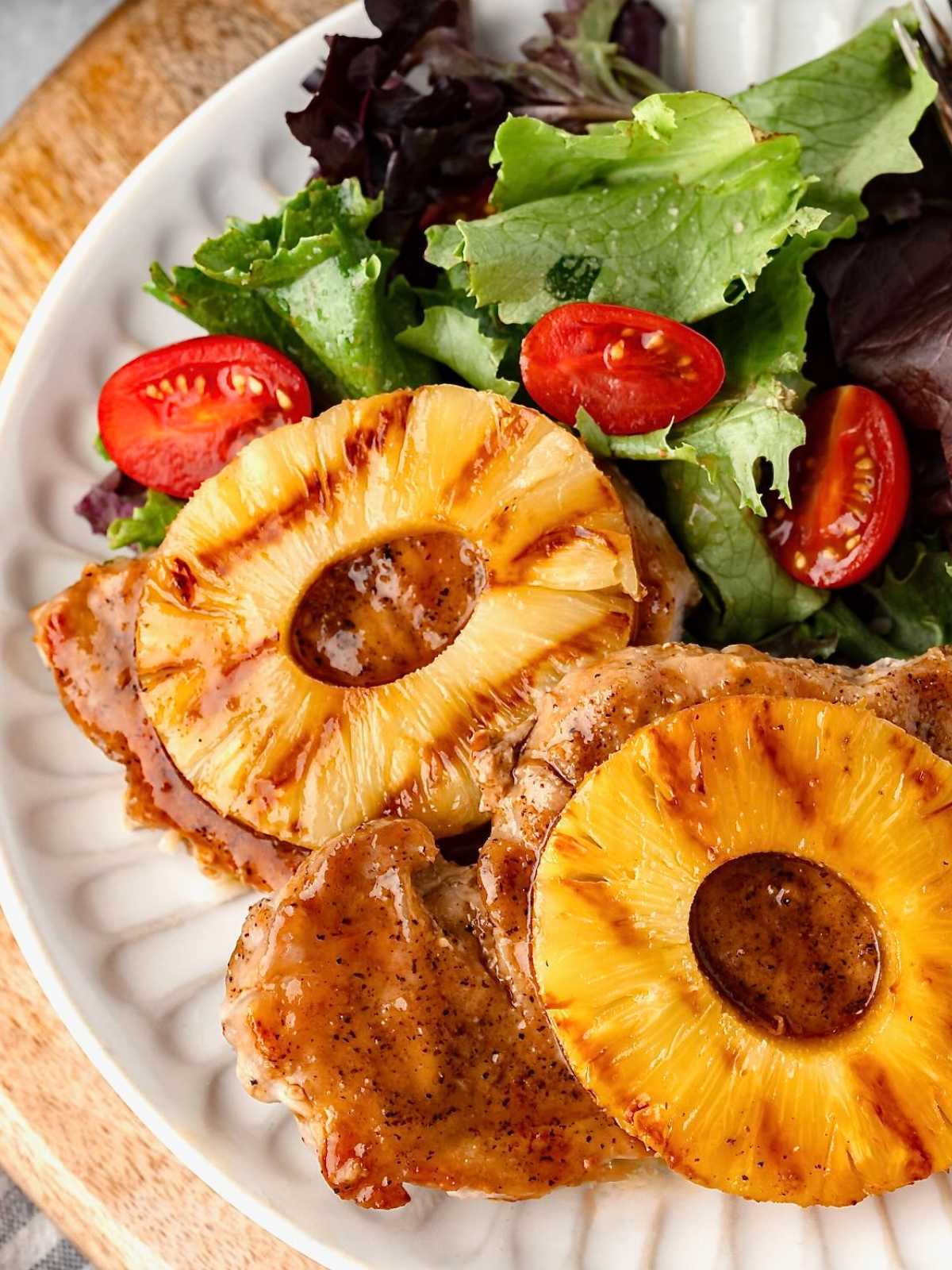 Grilled pork chops topped with pineapple rings, served with a green salad and cherry tomatoes on a white plate.