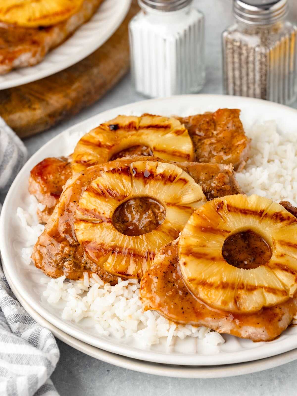 Pork chops topped with grilled pineapple rings served over white rice on a white plate.