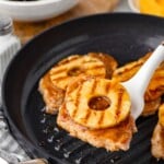A grilled pork chop topped with a pineapple ring being lifted from a skillet, with salad in the background.