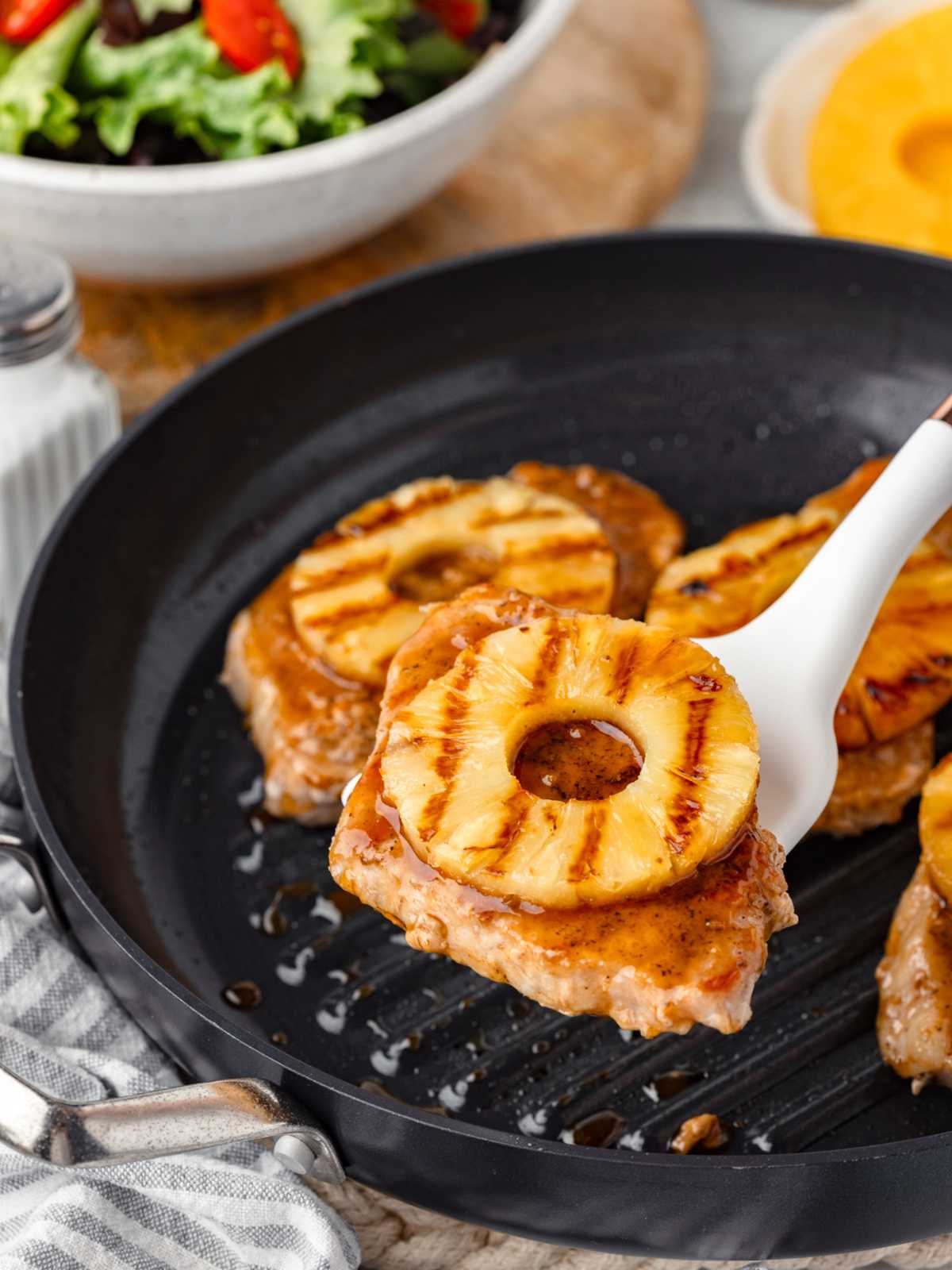 A grilled pork chop topped with a pineapple ring being lifted from a skillet, with salad in the background.