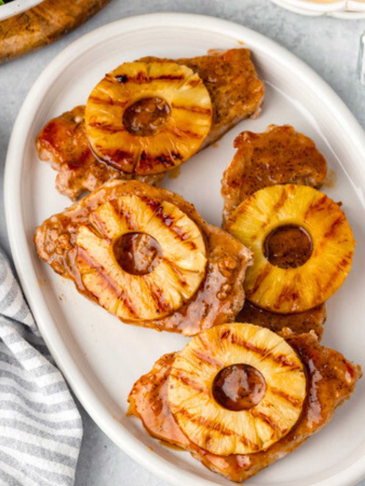 Four glazed pork chops topped with grilled pineapple slices on a white oval plate.