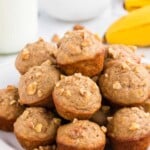 A plate of banana nut muffins topped with chopped walnuts, with bananas and a bowl of walnuts in the background.