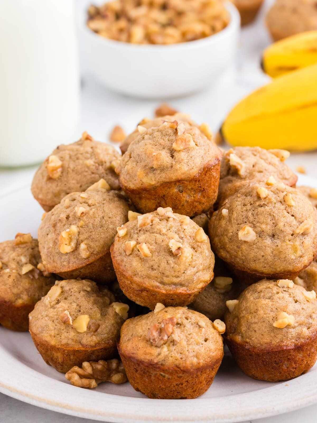 A plate of banana nut muffins topped with chopped walnuts, with bananas and a bowl of walnuts in the background.