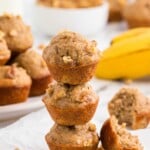 Three mini banana nut muffins stacked, with bananas and more muffins in the background. One muffin is broken open.