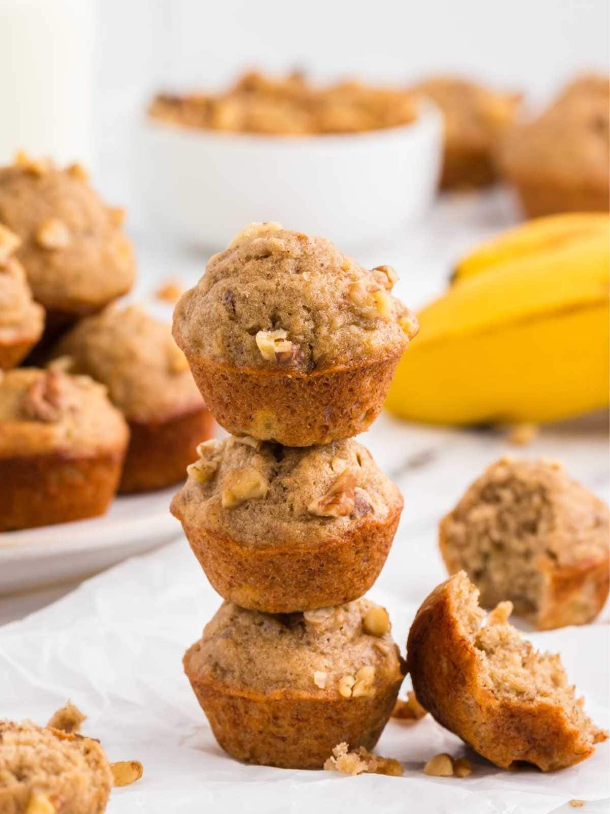 Three mini banana nut muffins stacked, with bananas and more muffins in the background. One muffin is broken open.