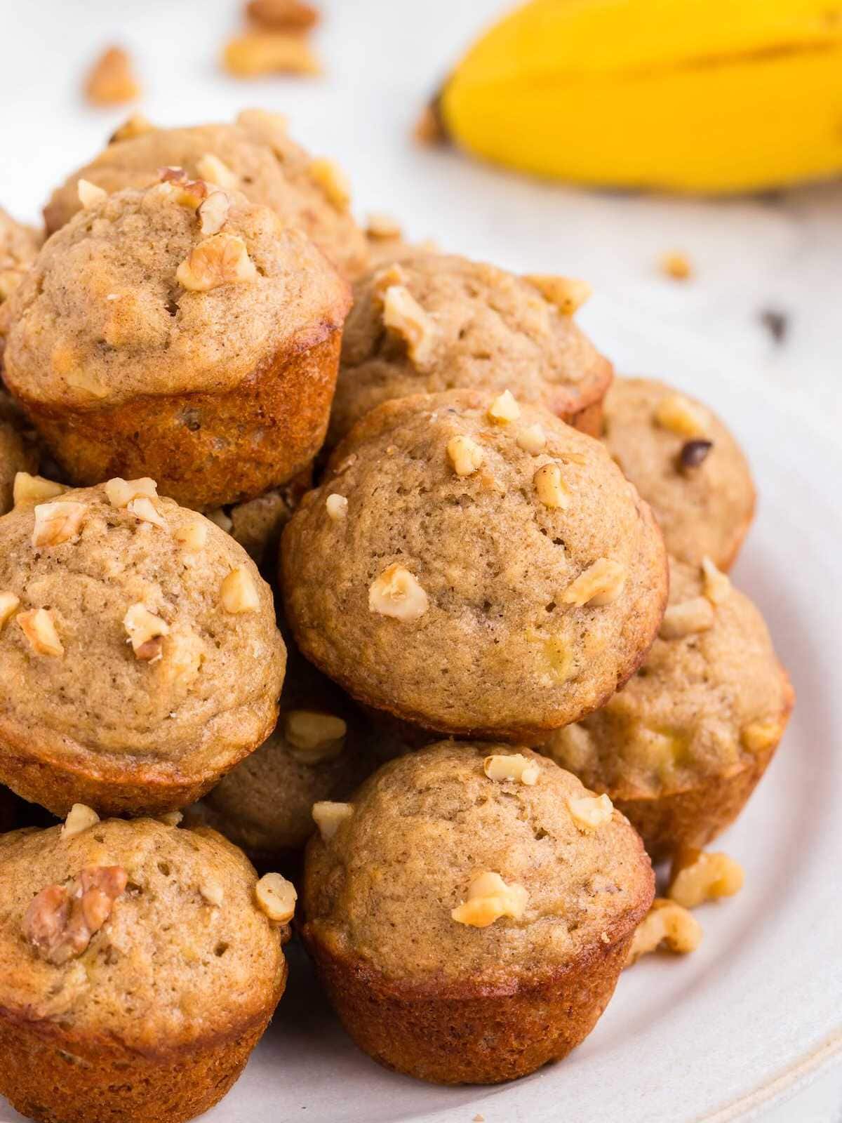 A plate of banana nut muffins topped with chopped walnuts, with a banana in the background.