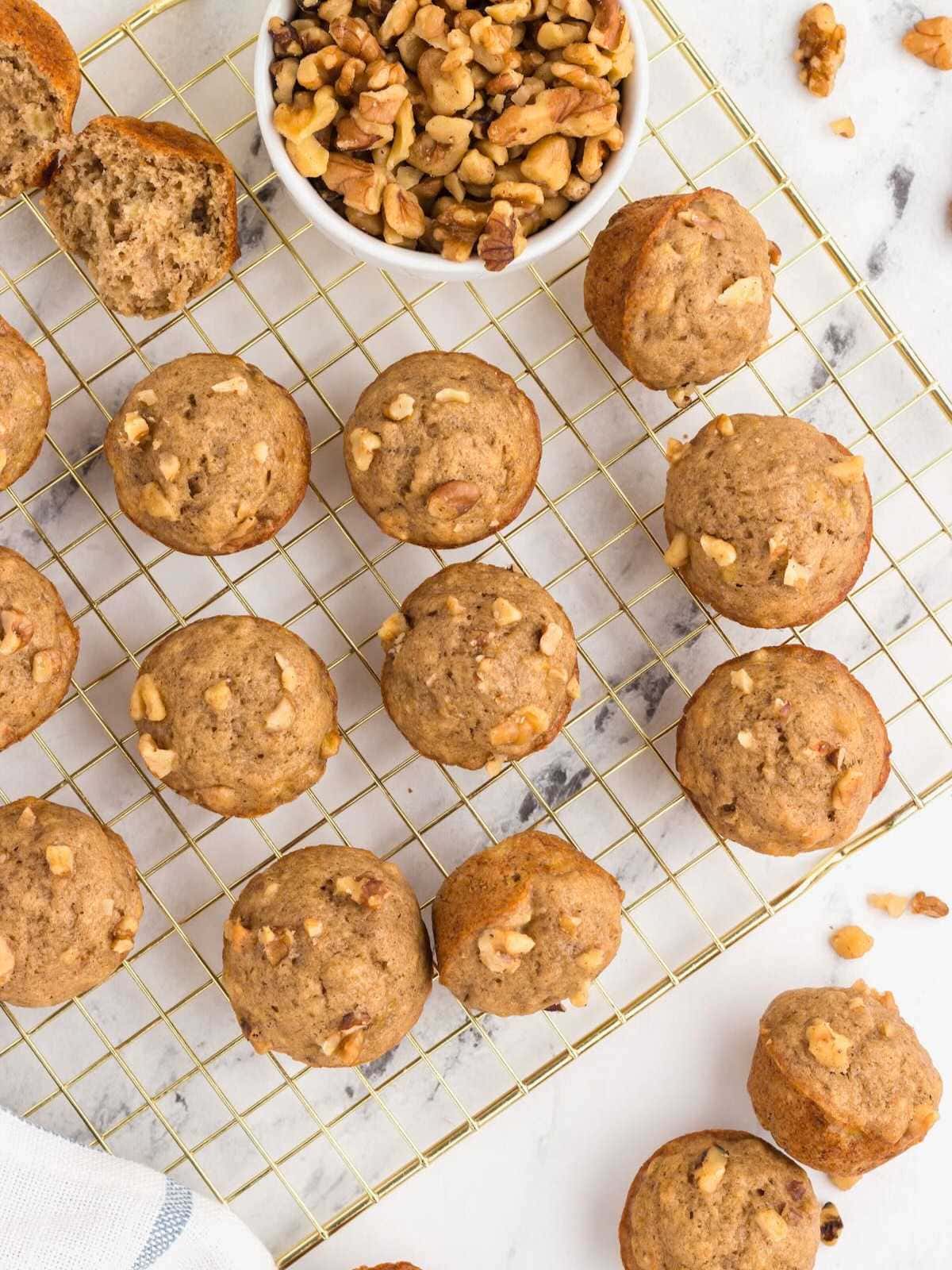 Mini banana nut muffins on a cooling rack with a bowl of chopped walnuts on a marble surface.