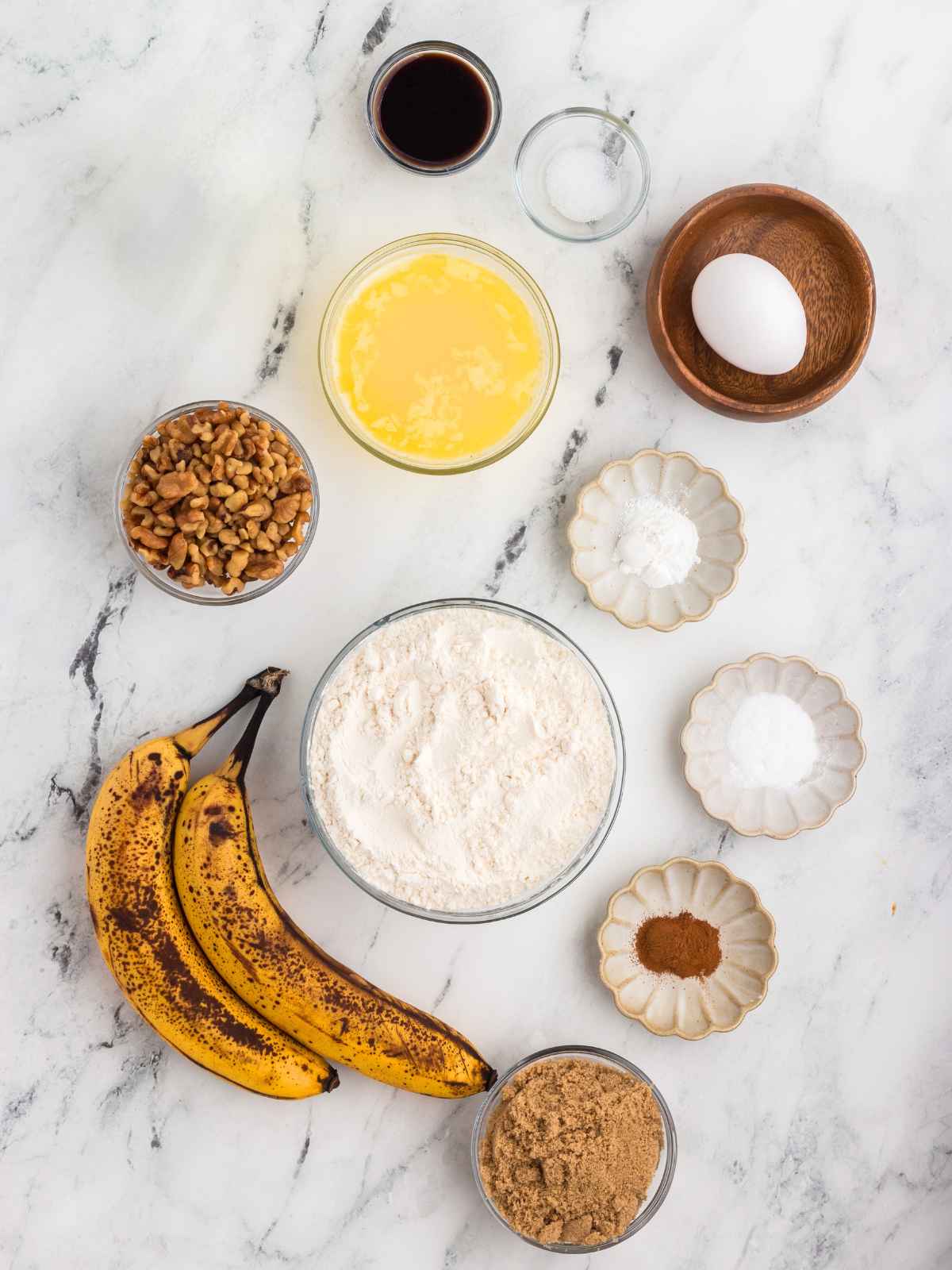 Several baking ingredients on a marble surface, including bananas, flour, egg, walnuts, butter, and spices.