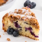 A slice of blueberry coffee cake with streusel topping and icing, served with fresh blueberries.