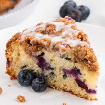 A slice of blueberry coffee cake with streusel topping and icing, served with fresh blueberries.