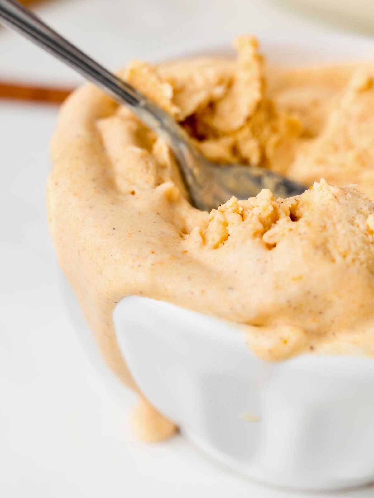 Creamy pumpkin ice cream overflowing from a white bowl with a spoon scooping some out.