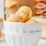 Three scoops of light brown ice cream in a stacked white bowl on a table, background softly blurred.