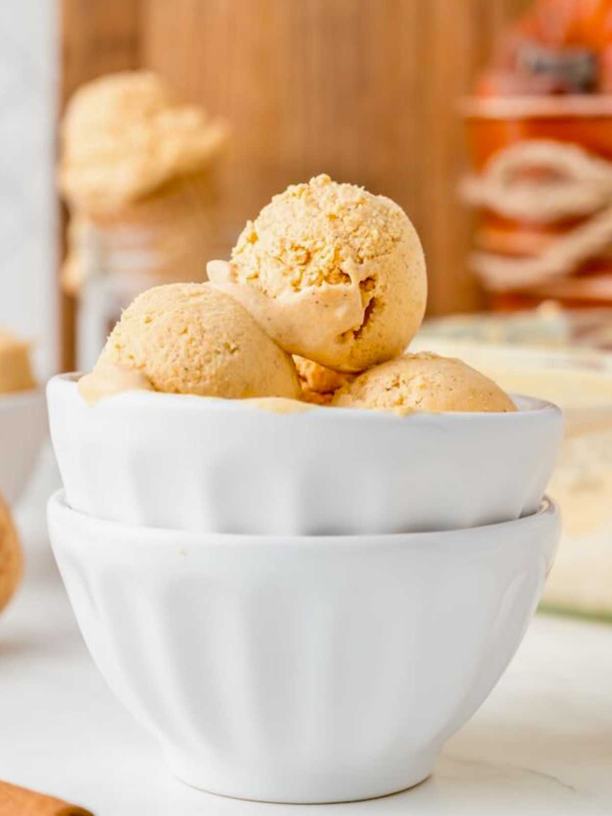 Three scoops of light brown ice cream in a stacked white bowl on a table, background softly blurred.