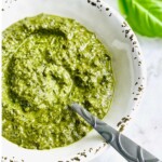 A bowl of green pesto sauce with a spoon, garnished with a basil leaf in the background.