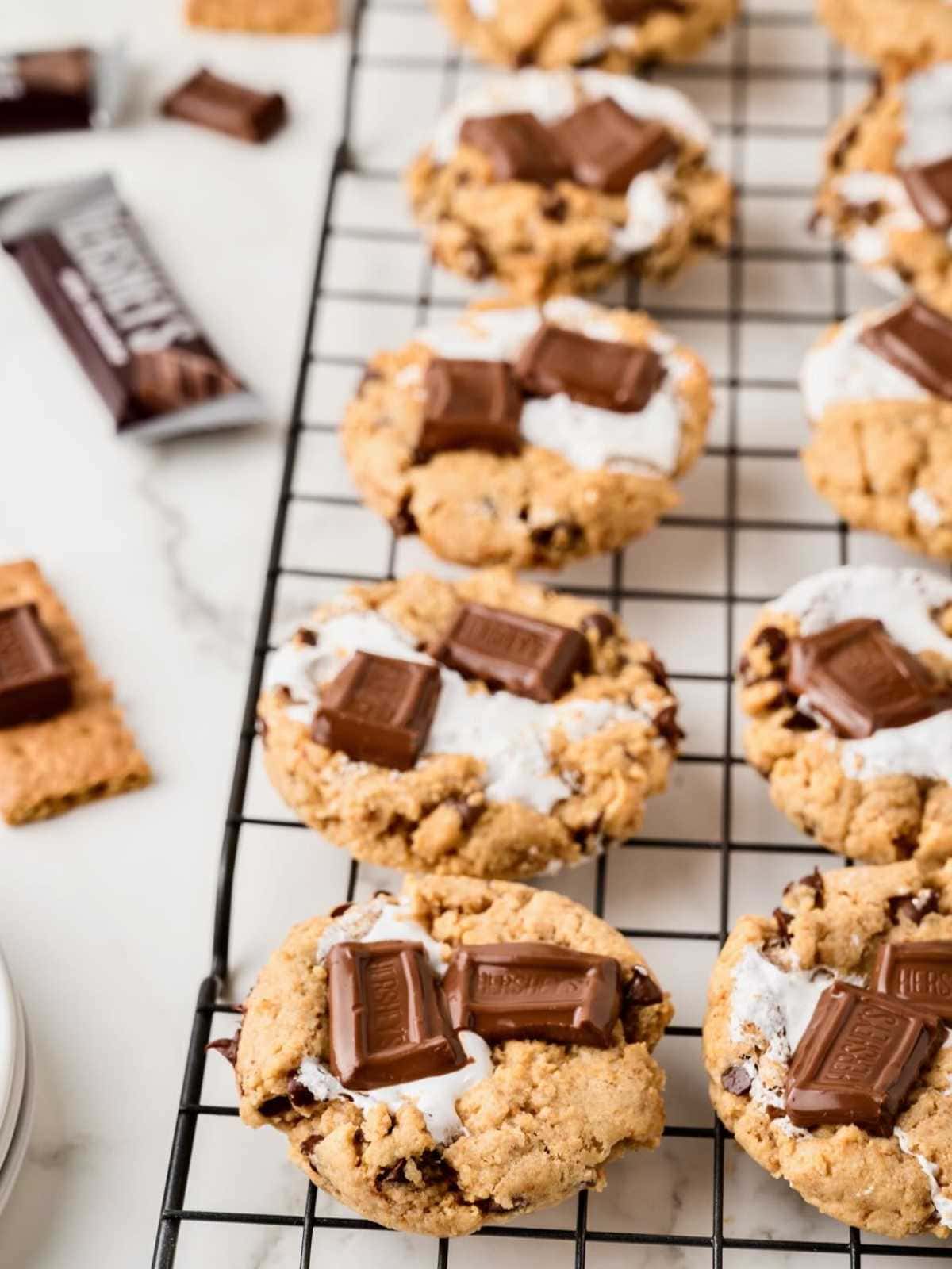 Cookies with melted marshmallow and chocolate pieces cooling on a wire rack, with graham crackers nearby.