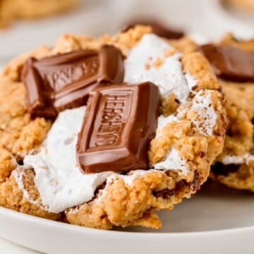 A cookie topped with melted marshmallow and Hershey’s chocolate pieces on a white plate.