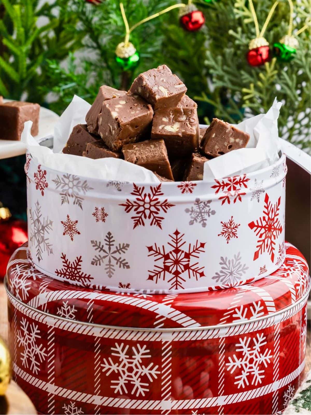A snowflake tin filled with chocolate fudge sits in front of a decorated Christmas tree.