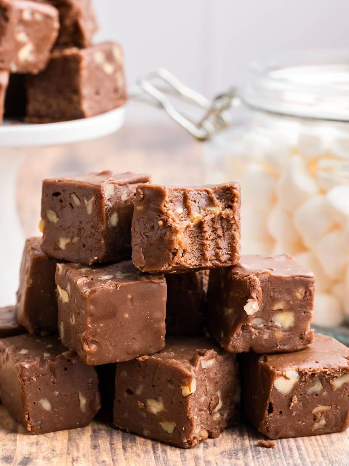 A stack of chocolate fudge pieces with nuts, one with a bite taken out, next to a jar of marshmallows.