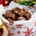 Chocolate fudge pieces stacked in a festive tin with red snowflake designs, surrounded by holiday decor.