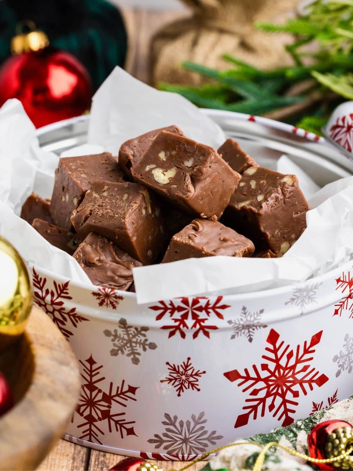 Chocolate fudge pieces stacked in a festive tin with red snowflake designs, surrounded by holiday decor.