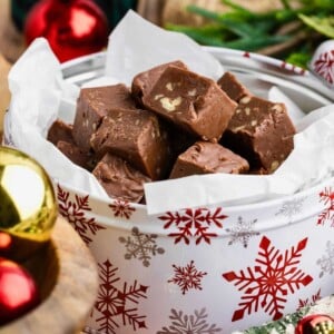 Chocolate fudge pieces with nuts in a festive tin decorated with red snowflakes, surrounded by ornaments.