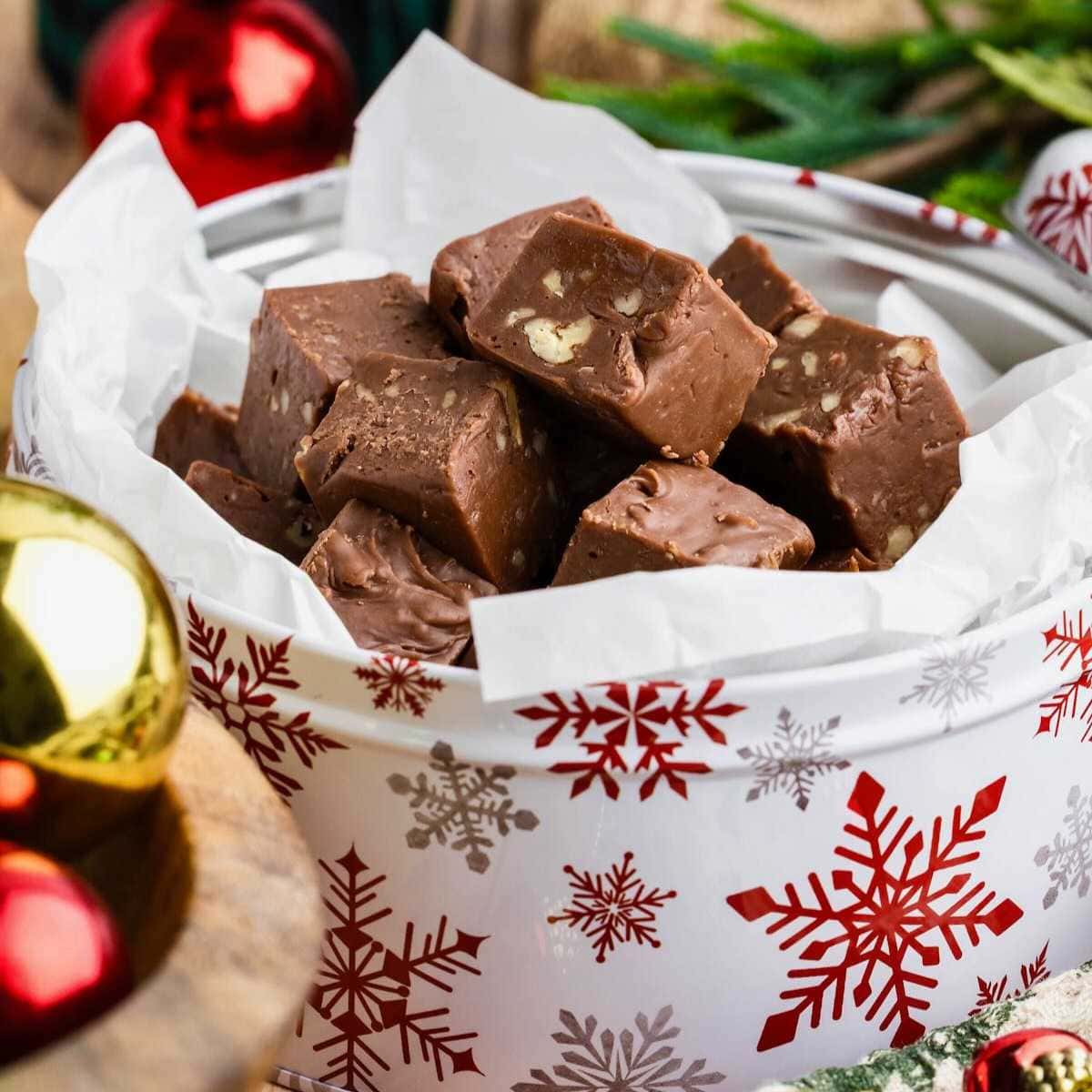 Chocolate fudge pieces with nuts in a festive tin decorated with red snowflakes, surrounded by ornaments.