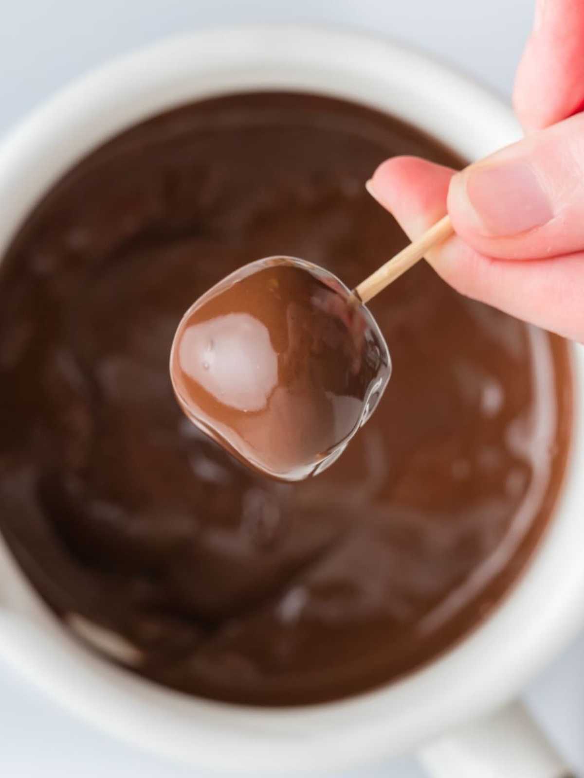 A hand holding a toothpick with a peanut butter ball dipped in melted chocolate over a bowl.